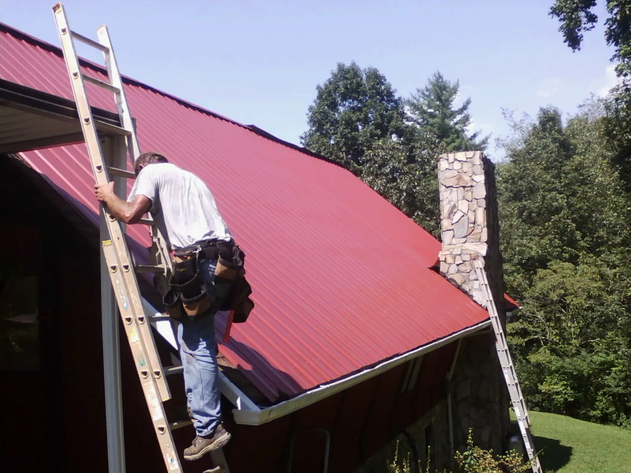 Roofer on a ladder beside a red metal roof and stone chimney — Soffit & Fascia Repair by Roofing Brothers in Middleton